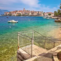 Widok na turkusowe morze i kamienistą plażę z kamiennymi schodami prowadzącymi do wody. W oddali widać urokliwe miasto Korcula, pełne zabytków i atrakcji.