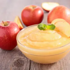 Un tazón de deliciosa salsa de manzana casera, decorado con menta y rodajas de manzana, rodeado de manzanas rojas frescas sobre una mesa de madera.