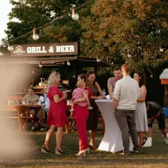 Ludzie rozmawiający przy stolikach i budce z napisem "Grill & Beer" na zewnątrz.