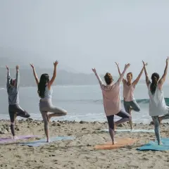 Fem kvinnor utför pilates yoga på stranden med havet i bakgrunden.