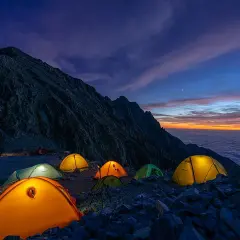 Acampada en la montaña al amanecer. Tiendas iluminadas sobre rocas, con un mar de nubes y cielo anaranjado. ¡Espero que no haya multa por acampar aquí!