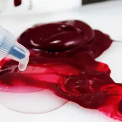 Un beccuccio trasparente rilascia un liquido chiaro su una macchia di vernice acrilica color bordeaux, creando sfumature vivaci.