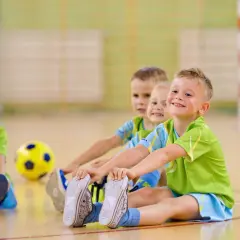 Trzech chłopców w zielonych koszulkach siedzi na podłodze sali gimnastycznej, obok żółtej piłki.