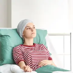 Młoda kobieta w chuście na głowie leży w szpitalnym łóżku, czekając na kolejną chemię. Czerwona w białe paski koszulka kontrastuje z zielonymi poduszkami.