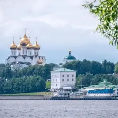 Złote kopuły cerkwi górują nad zielenią drzew nad brzegiem rzeki Wołgi. W oddali widać białe budynki i zielone dachy.