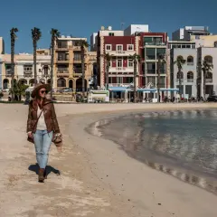 Kobieta w kapeluszu spaceruje po piaszczystej plaży z kolorowymi budynkami w tle.