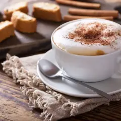 Un cappuccino senza latte, con schiuma soffice e cacao, accompagnato da biscotti secchi e cannella su un tavolo di legno rustico.
