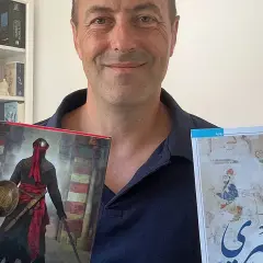 Juan de Granada, sonriente, sostiene dos libros. Uno muestra un guerrero con armadura y el otro, una escena histórica.