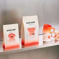 Dwie paczki kawy Coffee Plant z etykietami "Low Jungle Fruit" i "Low Very Berry" sugerują, jaka kawa ma najmniej kofeiny. Obok kubki.