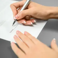 Mano femenina con uñas cuidadas escribe en un documento. El texto "Signature" es visible, pero el **contrato sin firmar** carece de **validez** hasta que se complete.