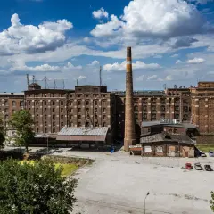 Dawny kompleks fabryczny z ceglanymi budynkami i wysokim kominem. Na placu przed budynkami zaparkowane samochody, a w tle zielone drzewa i błękitne niebo z chmurami. To miejsce, gdzie kiedyś tętniło życie, a dziś jest jak psie pole.