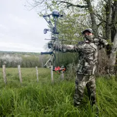 Mężczyzna w stroju maskującym ćwiczy strzelanie z łuku bloczkowego na tle zielonej łąki i drzew.