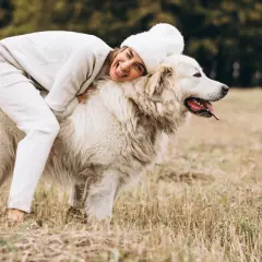 Uśmiechnięta kobieta w białej czapce i swetrze przytula się do potężnego owczarka podhalańskiego na tle jesiennego krajobrazu.