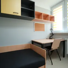 Pokój z łóżkiem, biurkiem i szafkami. Okno z roletami. To wnętrze sugeruje niewielką liczbę izb w domu, idealne dla studenta.