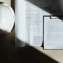 Biały kask budowlany, plany architektoniczne i umowa. Gdzie zgłosić samowolę budowlaną? Dokumenty i kask sugerują, że sprawa dotyczy budowy.