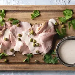 Fette di arrosto con salsa tonnata, capperi e prezzemolo su tagliere di legno.