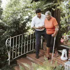 Ein Mann hilft einer älteren Frau mit Krücken die Treppe hinunter. Ihr Gang wirkt unsicher, aber sie lächelt.