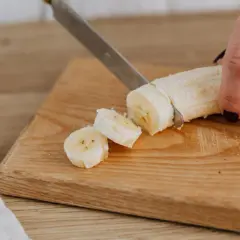 Eine Hand schneidet eine Banane in Scheiben auf einem Holzbrett. Perfekte Energie für das Training.