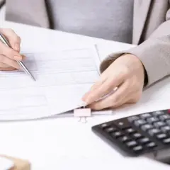 Kobieta analizuje dokumenty, liczy koszty eksploatacyjne w spółdzielni. Obok kalkulator.