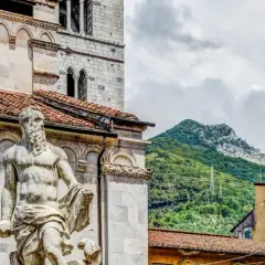 Statua nel carrara centro storico, con montagne verdi sullo sfondo e case colorate.