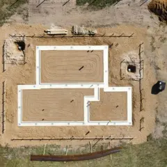 Widok z lotu ptaka na budowę domu. Widać fundamenty pośrednie, piasek, wykopy i materiały budowlane.