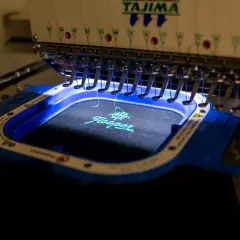 Machine à broder Tajima créant une estampille digitalisée sur un tissu noir. Le logo "Hooper" est en cours de réalisation avec du fil turquoise.