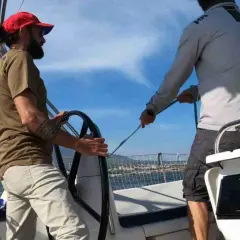 Due uomini su una barca a vela sport, uno al timone, l'altro che regola le cime. Cielo azzurro e mare calmo.