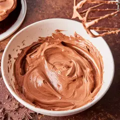 Schoko-Buttercreme in einer Schüssel, bereit zum Verzieren eines Kuchens. Ein Handmixer mit Schoko-Buttercreme hängt darüber.
