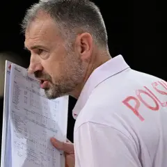 Trener Nikola Grbić wpatrzony w dokumenty, z napisem "POLSKA" na koszulce. Może analizuje statystyki, myśląc o swojej żonie.