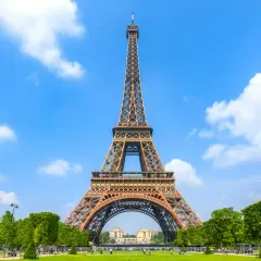 The Eiffel Tower stands tall against a bright blue sky with fluffy white clouds, surrounded by lush green trees and a manicured lawn.