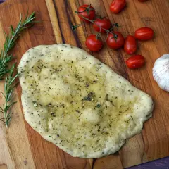 Ein unfertiges Pizzabrot mit Knoblauch und Kräutern liegt auf einem Holzbrett, daneben Kirschtomaten und Rosmarin.