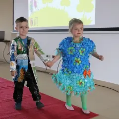 Dzieci prezentują stroje ekologiczne. Dziewczynka w niebieskiej sukience z nakrętek, chłopiec w kurtce z opakowań. Inspiracja, jak zrobić strój ekologiczny dla dziewczynki.