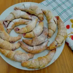 Pyszne, domowe rogaliki z marmoladą, posypane cukrem pudrem, ułożone na talerzu obok ręcznika z truskawkami.