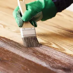 Mano con guante verde aplicando barniz sobre madera, creando un acabado brillante. Barnizar sobre barniz sin lijar para renovar la superficie.