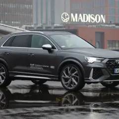 Szary Audi RS Q3 na mokrej nawierzchni, z odbiciem w kałuży. W tle budynek z napisem MADISON.