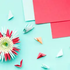 Pyssel med papper: en färgglad 3D-origamiblomma omgiven av små vikta pappersdelar och ark i rött och ljusblått.