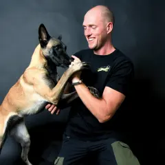Ein Mann lacht und hält die Pfoten seines Malinois. Erfolgreiches Hunde Training zeigt die enge Bindung.