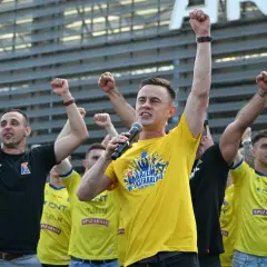 Mężczyzna w żółtej koszulce z mikrofonem i kibice z podniesionymi pięściami.