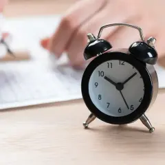 Czarny budzik na drewnianym blacie, w tle ręce piszące na arkuszu.