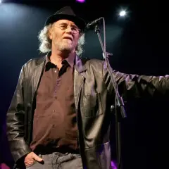 Francesco De Gregori, con cappello e giacca di pelle, canta sul palco. I suoi testi, come "Adelante Adelante", risuonano nell'aria.