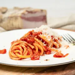Ein Teller Pasta all'Amatriciana, garniert mit geriebenem Parmesan. Im Hintergrund ein Stück Guanciale, die Zutat für das authentische Amatriciana Rezept.