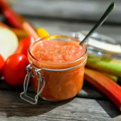 En burk med rabarber chutney står på ett träbord, omgiven av färska rabarber, tomater och en lök.