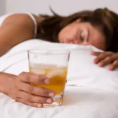 Kobieta śpi w łóżku, trzymając szklankę z napojem i lodem. Może to być alkohol, np. GHB.