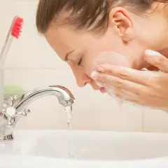 Kobieta myje twarz w umywalce, zastanawiając się, ile razy dziennie myć twarz. Obok stoją szczoteczki do zębów.