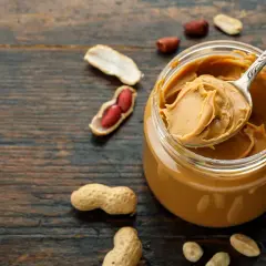Ein Löffel cremiger Erdnussbutter aus einem Glas, umgeben von Erdnüssen. Erdnussbutter gesund und lecker!