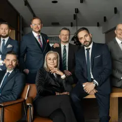 Grupa profesjonalistów w garniturach, siedząca i stojąca, w nowoczesnym wnętrzu.
