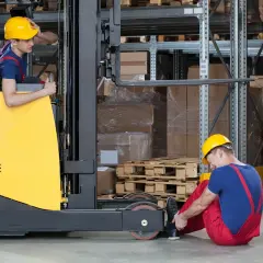 Ein Mann mit gelbem Helm hat sich am Fuß verletzt, als er neben einem Gabelstapler sitzt. Ein Kollege im Stapler scheint besorgt. Ein Stapler Unfall.
