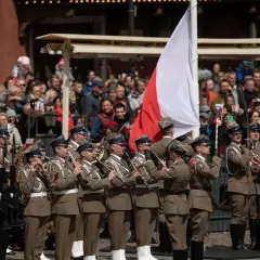 Orkiestra wojskowa grająca na instrumentach dętych na tle tłumu i polskiej flagi.