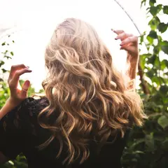 Blondes Haar mit Beach Waves im Sonnenlicht, umgeben von grünen Blättern.
