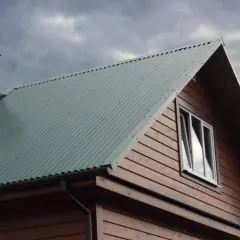 Dach domu pokryty zieloną blachą trapezową, widoczny komin i okno. Szerokość krycia blachy trapezowej zapewnia szczelność.